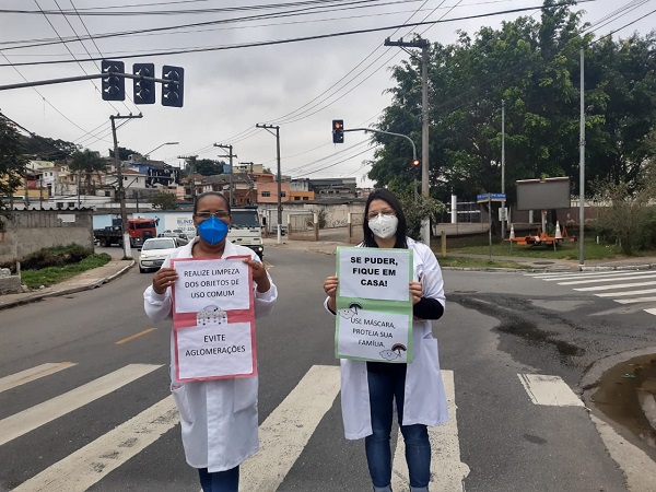 #PraCegoVer: Fotografia das profissionais da saúde realizando ações, todas as três estão em pé segurando cartazes de prevenção contra a COVID-19.